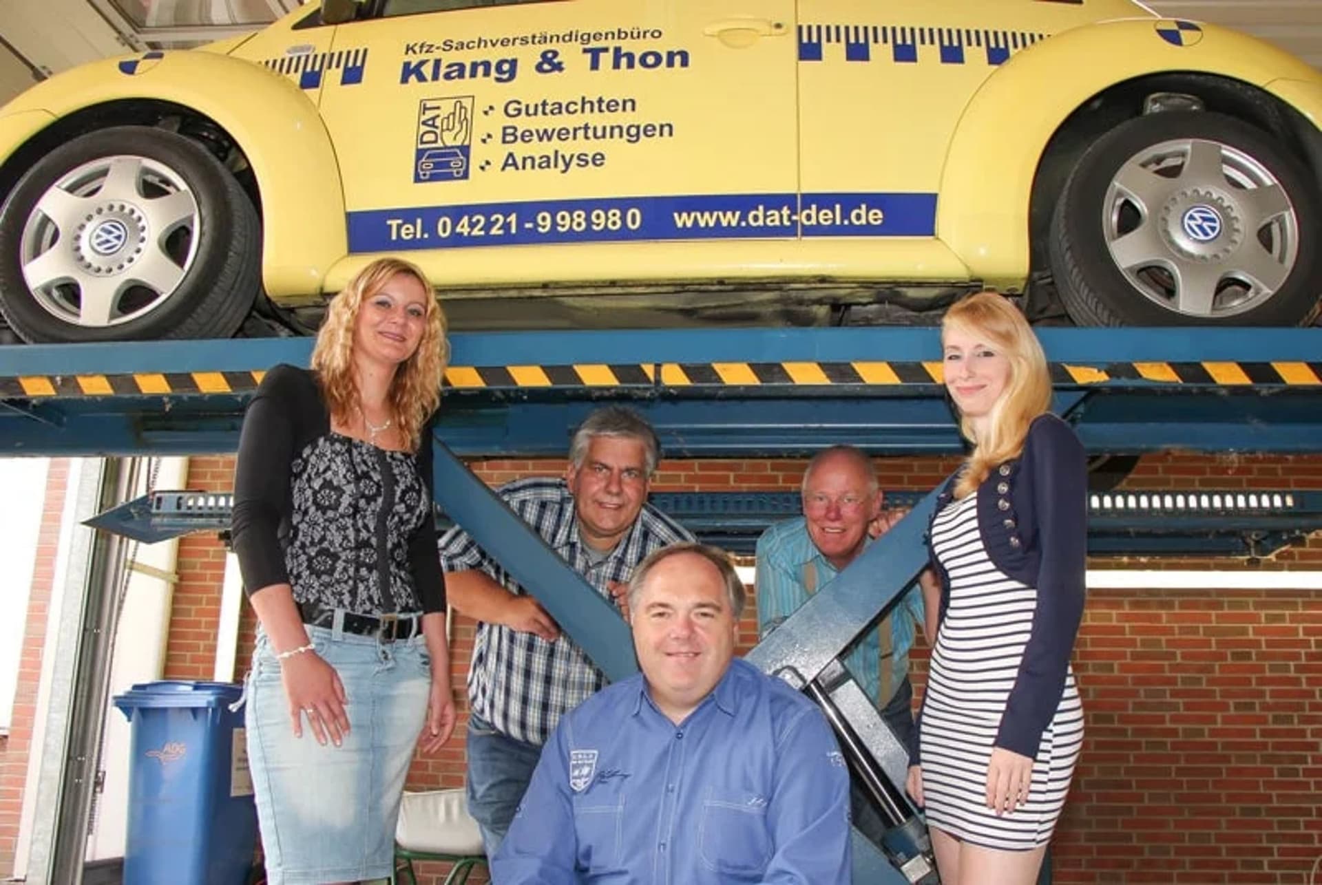 Fünf Personen vor einem gelben VW Käfer auf einer Hebebühne mit Firmenwerbung „Klang & Thon“.